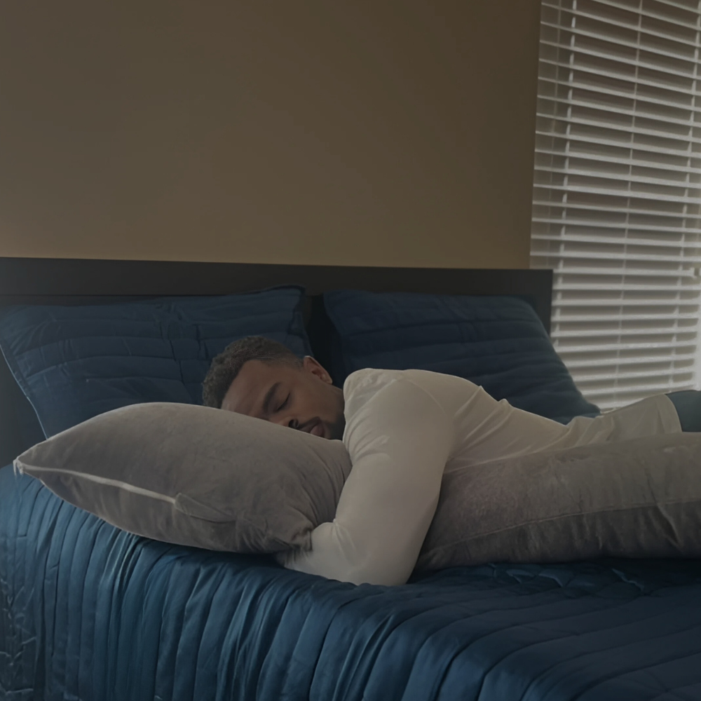 This image captures a peaceful moment of a man sleeping on his stomach, his head resting comfortably on the Special T Pillow. The neutral tones of the pillow and dark blue bedding, along with the soft morning light filtering through the blinds, evoke a serene sleeping environment, showcasing the pillow's comfort and support.