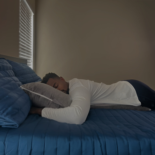 This image shows a man in a restful sleep while lying on his side, embracing the Special T Pillow. The room's muted lighting and soft color palette underscore the comfort and tranquility provided by the pillow, set against a rich blue bedspread.