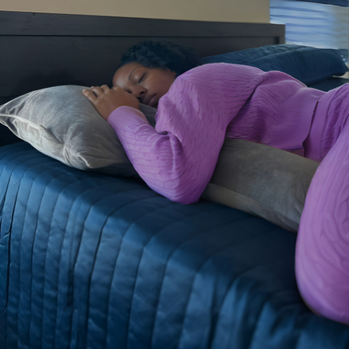 This image depicts a woman deeply asleep while hugging the Special T Pillow, dressed in a pink robe. The dark blue bedding and subdued room lighting enhance the sense of night-time comfort and the pillow's effectiveness in promoting restful sleep.