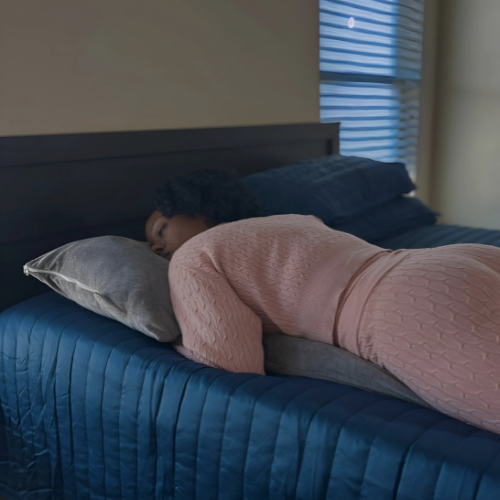 This image captures a serene scene of a woman sleeping on a Special T Pillow, showcasing the pillow's practical use. She is comfortably positioned on her stomach, highlighting the pillow's design that caters specifically to stomach sleepers. The calm, dimly lit bedroom setting emphasizes the pillow's role in promoting a restful sleep.